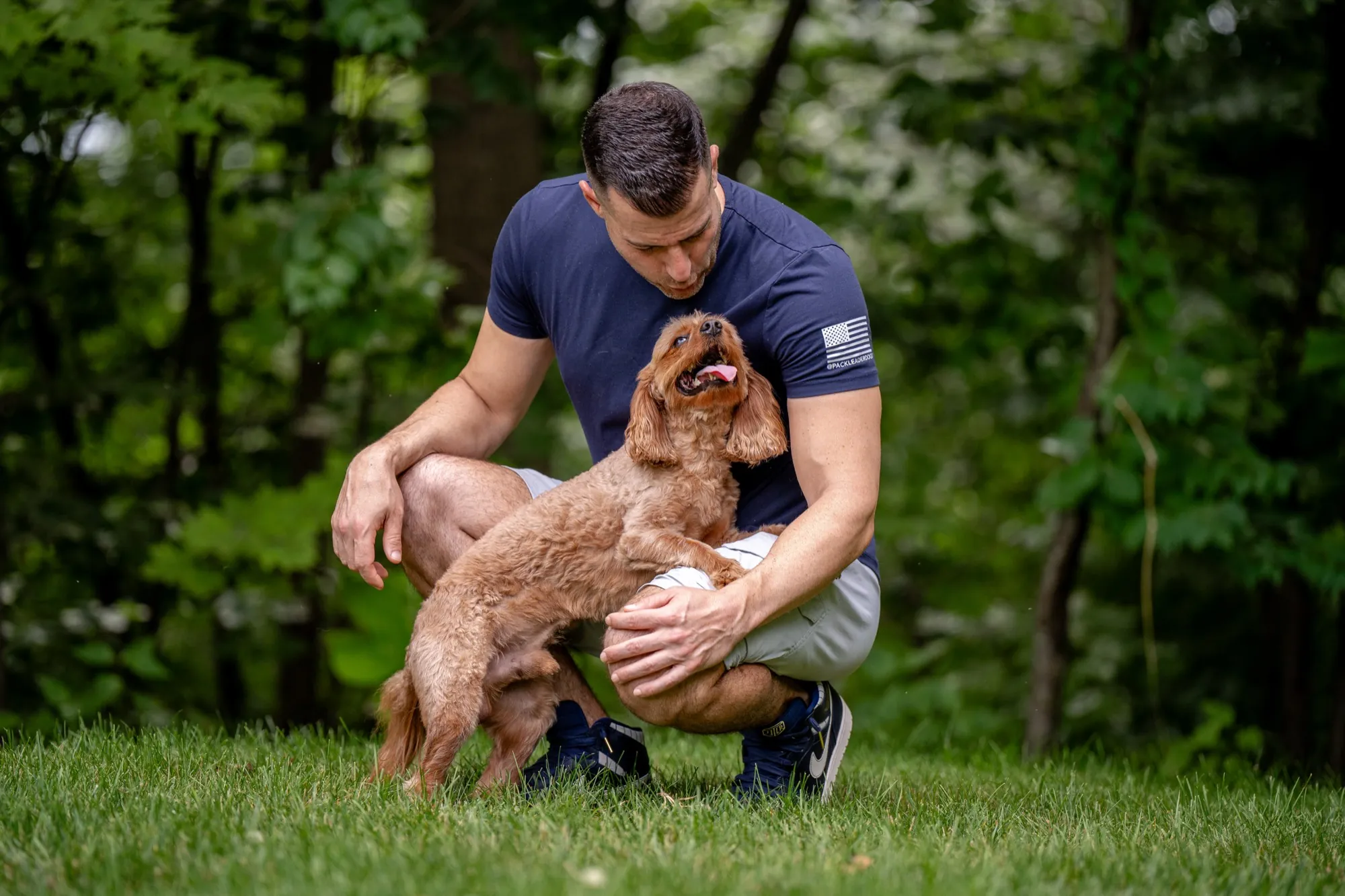 Steve Del Savio crouched on the lawn at The Maddie Ranch, a young cavapoo standing into his arms and looking up at him.