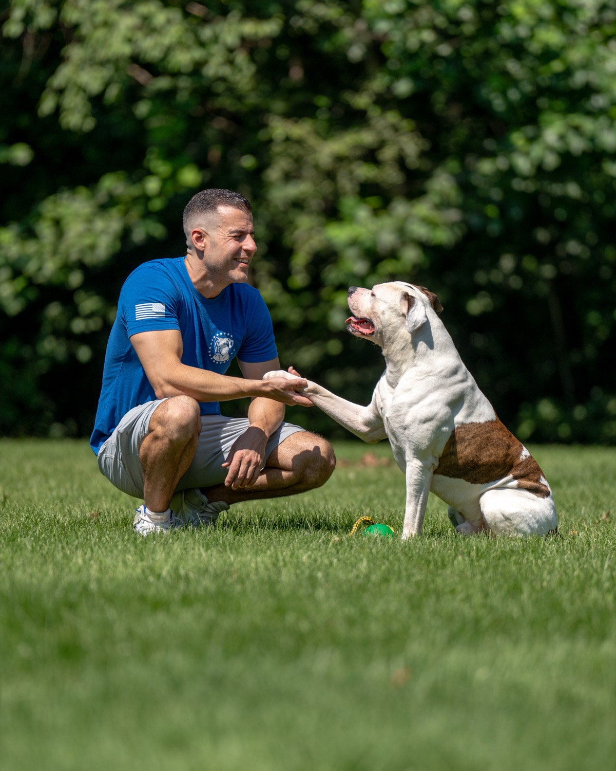 Steve Del Savio crouched on the lawn, smiling and holding the offered paw of a white and brindle American bulldog.