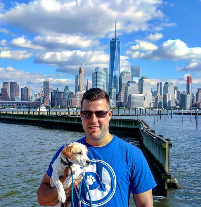 Cookie carried in the arms of a Pack Leader Dogs trainer on the waterfront, the Manhattan skyline behind them.
