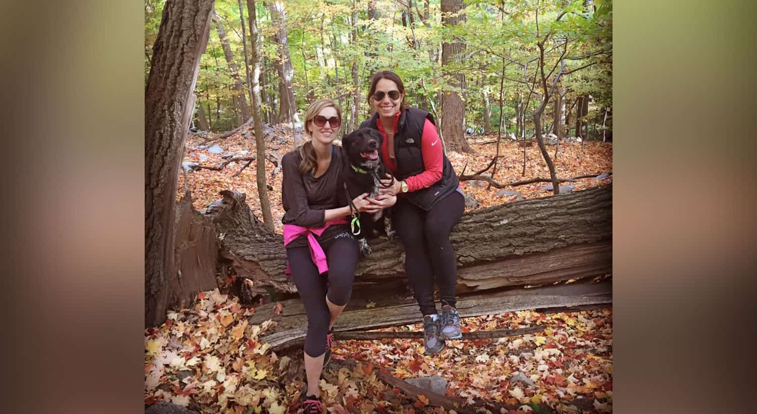 Rachel seated on a fallen log in the autumn woods, Marley the black dog resting between her and a friend.